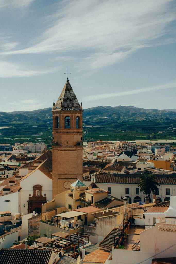 A tall church tower with a pointed roof overlooks a town with white buildings and distant mountains under a clear blue sky.