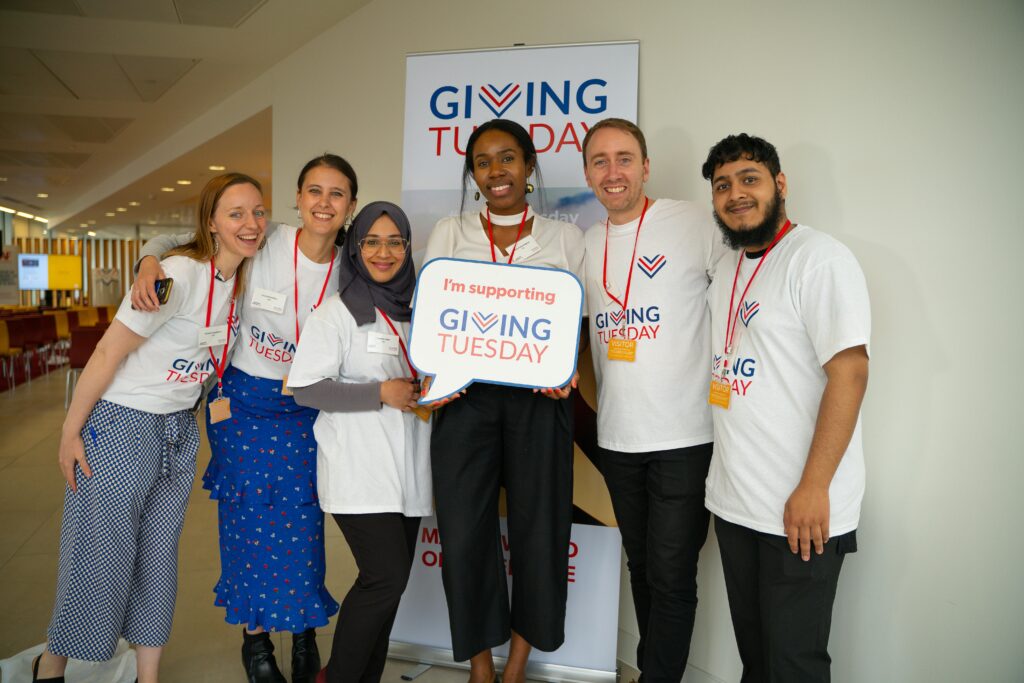 A group of six people wearing "Giving Tuesday" shirts stand together, smiling and holding a sign that says, "I'm supporting Giving Tuesday."