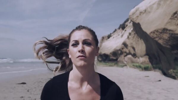 A woman with her hair blowing in the wind runs on a beach with large rocks in the background.