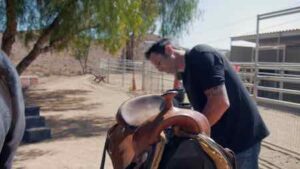 **Alt text:** A person is adjusting a saddle on a horse in an outdoor area with trees and fencing nearby.