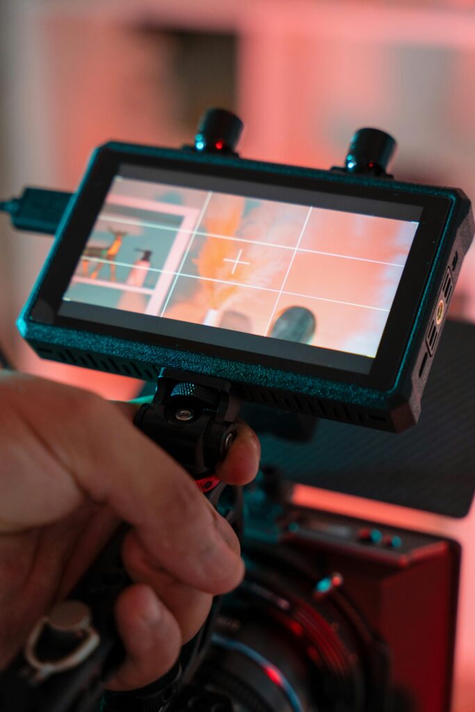 **Alt text:** A person holds a camera with an attached monitor displaying an image of a shelf with a vase and pampas grass, viewed through a crosshair grid.