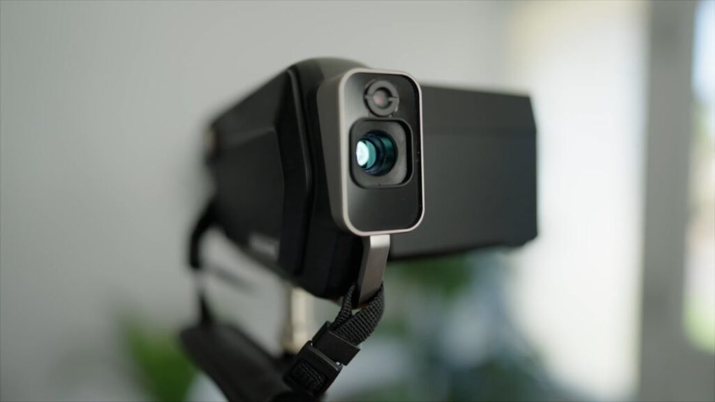 A close-up of a digital camera lens with a strap on a tripod, with a blurred background.