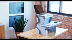 **Alt Text:** A wooden desk features an ergonomic stand holding a laptop at eye level, with a separate keyboard and mouse below, against a brick wall and near a plant by a window.