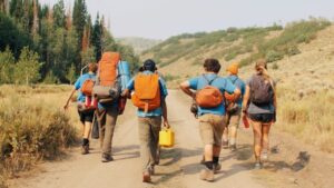 A group of people with backpacks walk on a dirt trail surrounded by trees and hills.