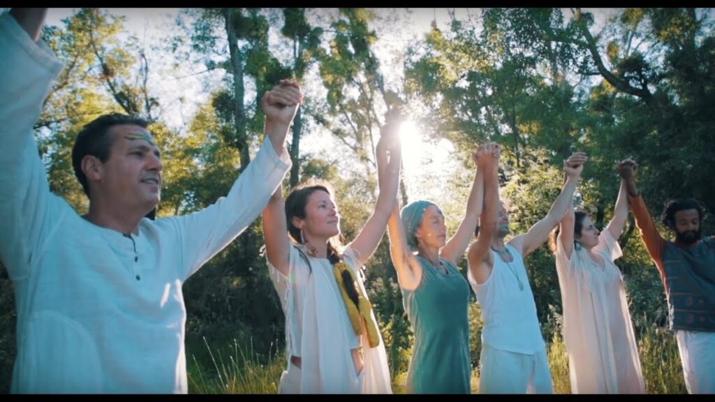 **Alt text:** A diverse group of people standing outdoors holding hands above their heads in a circle, surrounded by trees with sunlight streaming through.