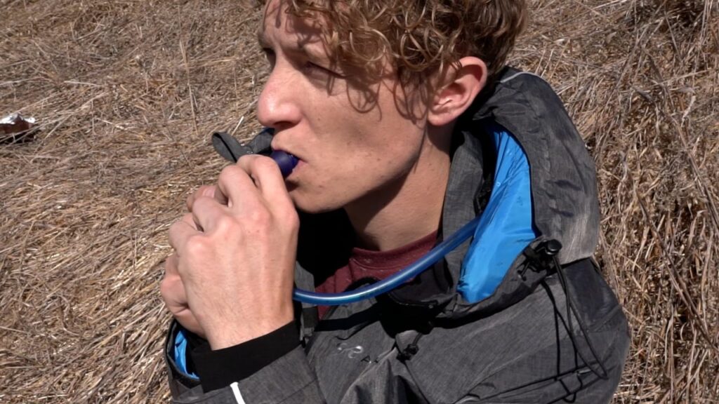 A person drinks from a hydration pack while sitting outdoors on dry grass.