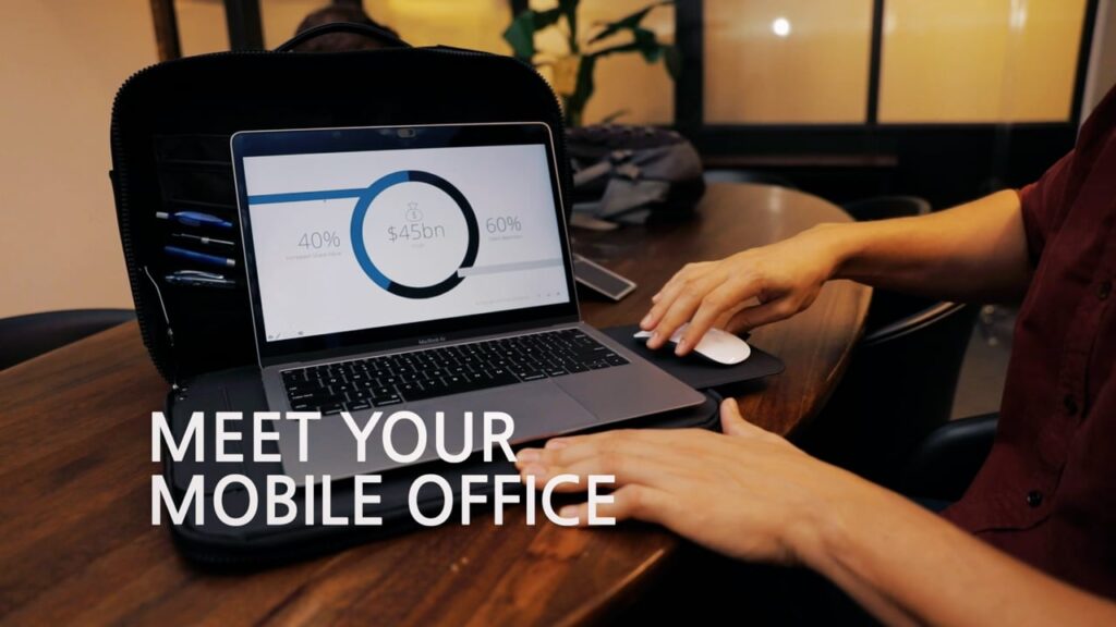 A person uses a laptop on a wooden table with a circular chart displayed on the screen and the text "MEET YOUR MOBILE OFFICE" visible beside it.