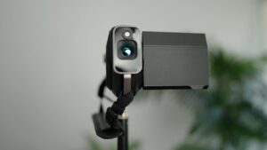 A digital camera mounted on a tripod with a blurred green plant in the background.