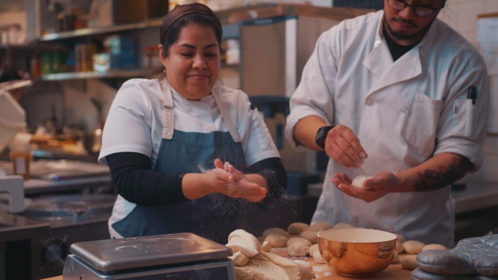 Two bakers in a kitchen preparing dough on a table with several pieces of dough and a copper bowl.