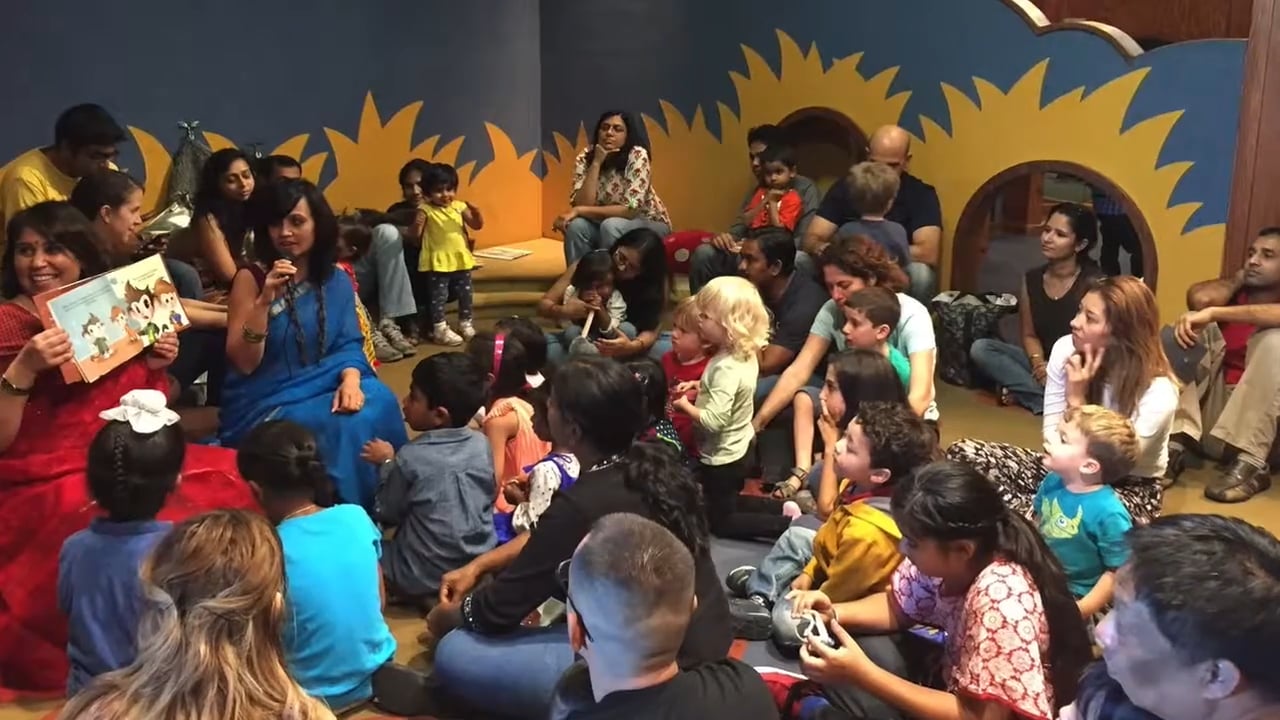 A group of children and parents sit on the floor, attentively listening to a woman in a blue dress holding a microphone during a storytime session.