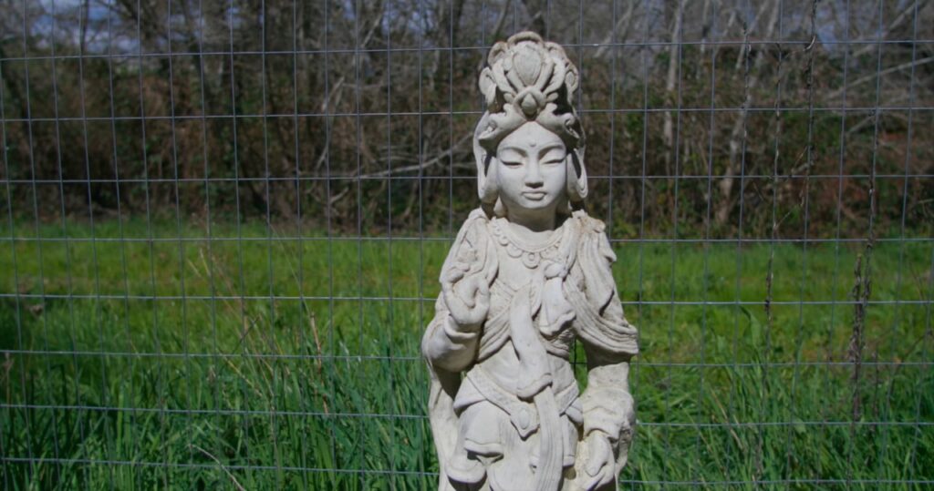 A stone statue of a serene figure with intricate robes stands in front of a wire fence, with green grass and trees in the background.