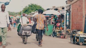 **Alt text:** A busy market scene with people walking and a man pushing a wheelbarrow along a path lined with stalls and items for sale.