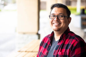 A person with glasses smiles while sitting at a wooden table, wearing a red and black checkered shirt.