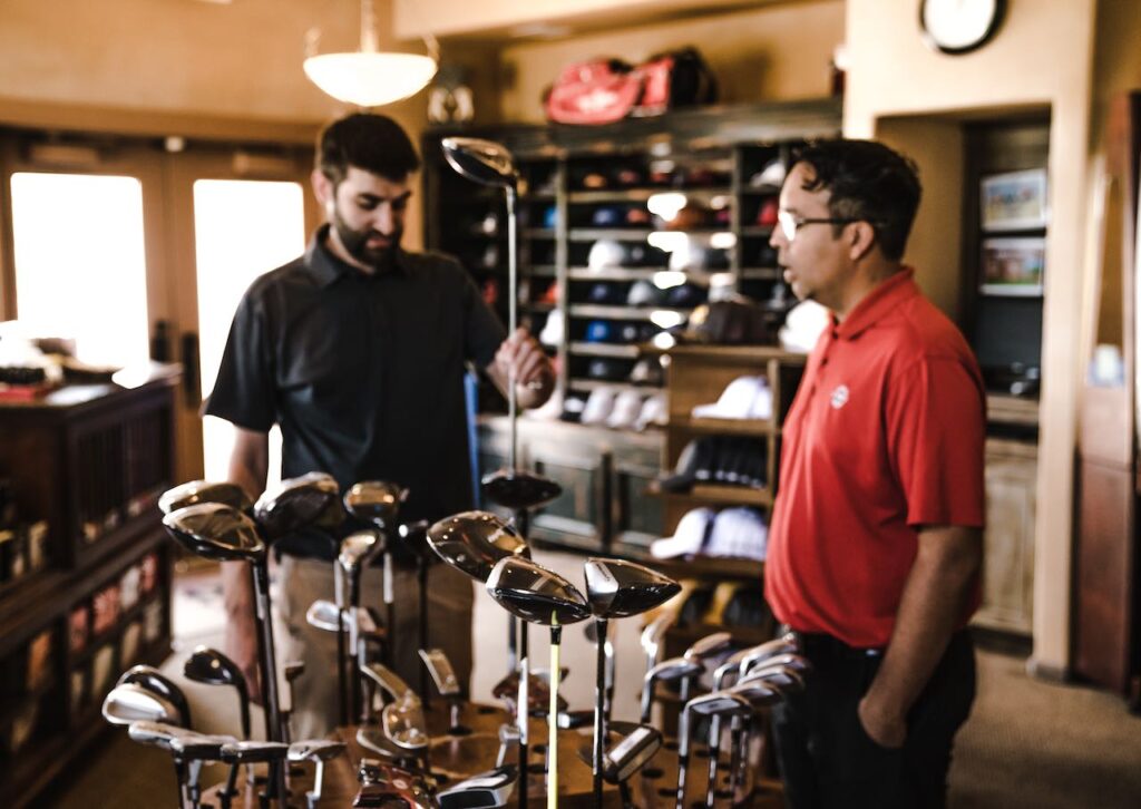 **Alt Text:** Two men are standing in a store examining a collection of golf clubs displayed on a table.