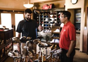 **Alt Text:** Two men in a golf shop examine various golf clubs displayed on a rack.
