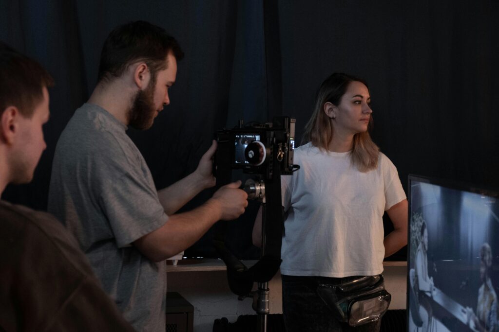 "A man operates a camera while a woman stands nearby in a dimly lit room with a television screen showing two people in conversation."