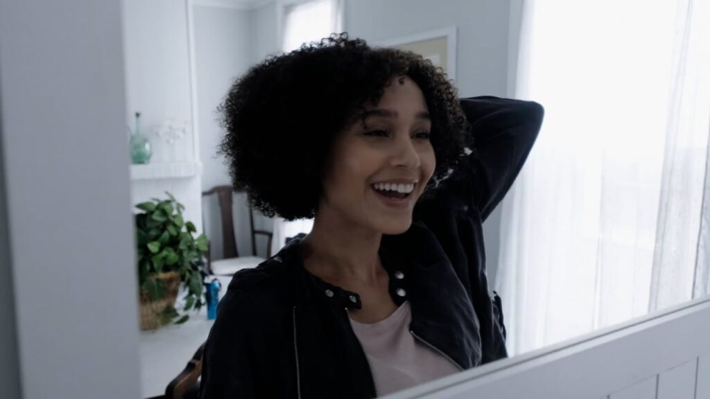 A person with curly hair smiles while looking at themselves in a mirror in a well-lit room.