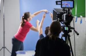 A woman is stretching to adjust objects against a blue background while a camera crew records the scene.