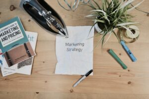 A crumpled paper labeled "Marketing Strategy" is placed on a wooden desk surrounded by books, an iron, markers, a plant, and a tape measure.