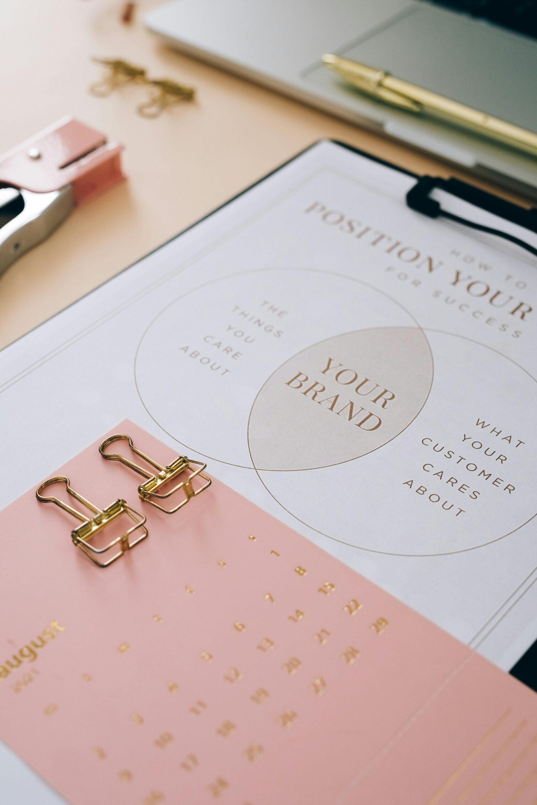 **Alt Text:** A desk setup with a pink calendar for August, two gold binder clips, and a chart on brand positioning overlapping interests between the brand and customers.