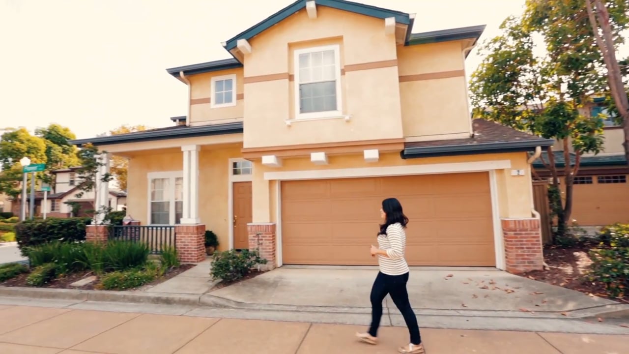 A woman walks in front of a two-story house with a beige exterior and a two-car garage.