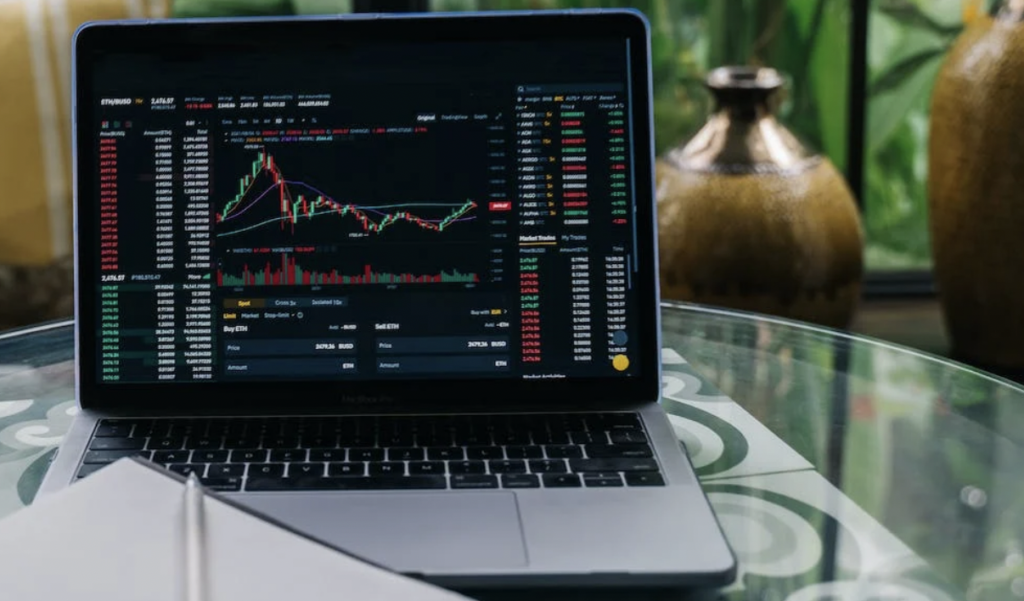 **Alt text:** A laptop on a glass table displaying cryptocurrency market charts and data.