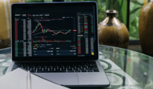 **Alt text:** A laptop on a glass table displaying cryptocurrency market charts and data.