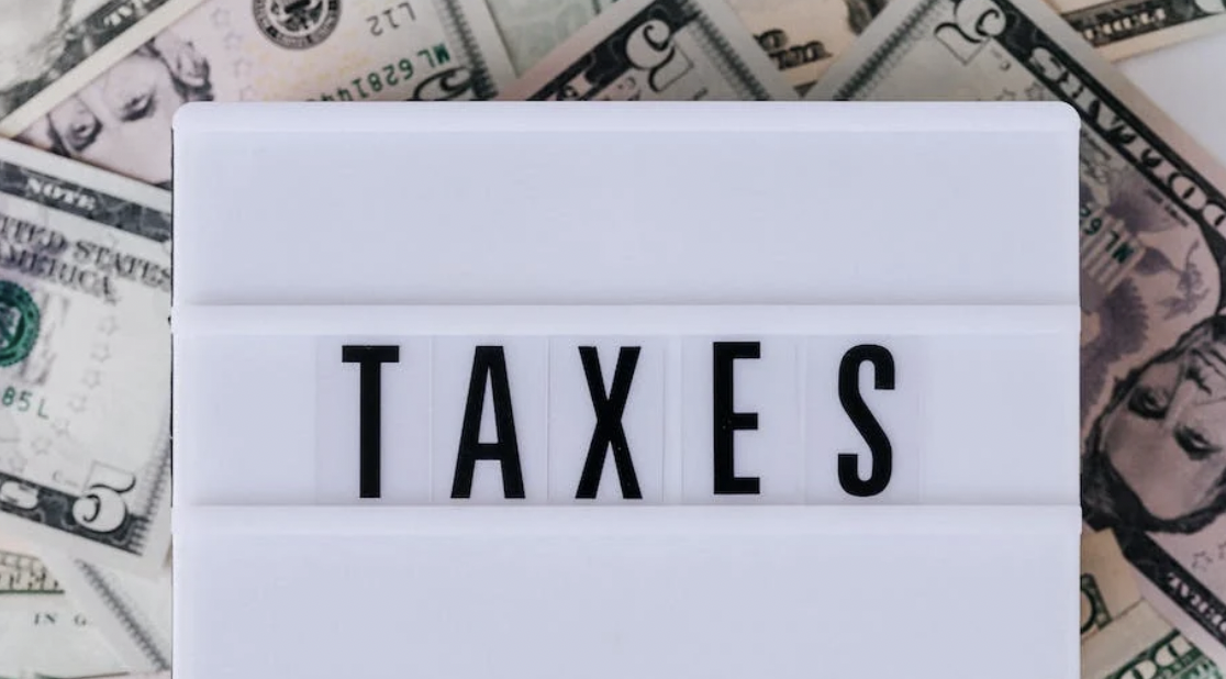 A sign displaying the word "TAXES" is placed on top of scattered U.S. dollar bills.