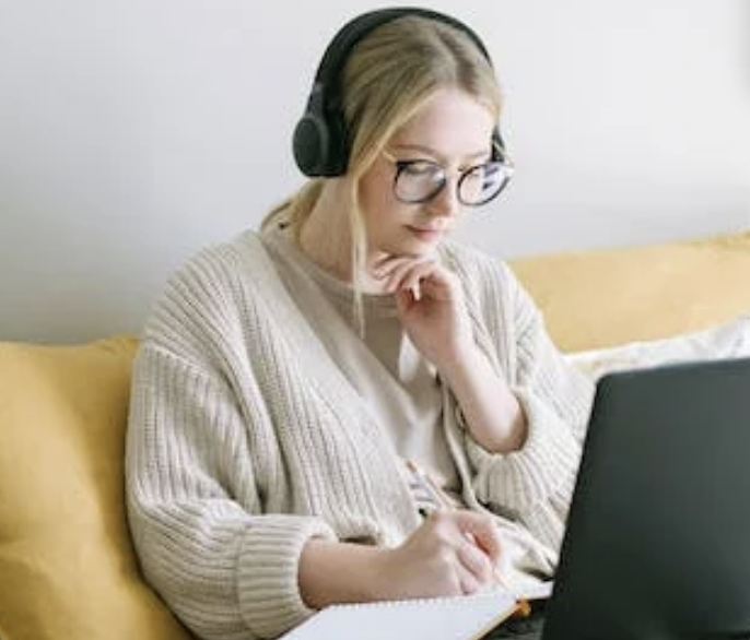 A person wearing headphones and glasses is sitting on a couch, focusing on a laptop and taking notes in a notebook.