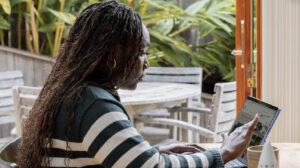A woman sitting outdoors using a laptop with greenery in the background.