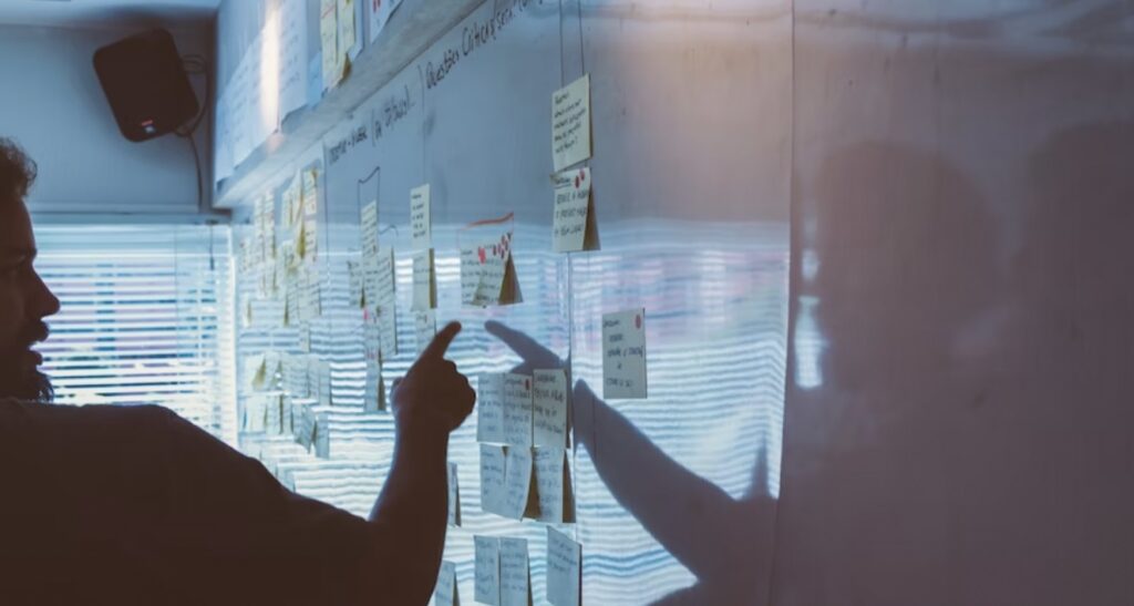 **Alt text:** A person points at sticky notes arranged on a large whiteboard with their shadow visible on the surface.