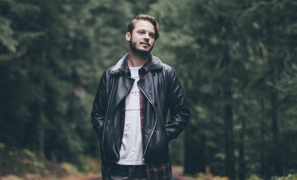 **Alt text:** A man wearing a leather jacket stands on a forest path, surrounded by tall trees.