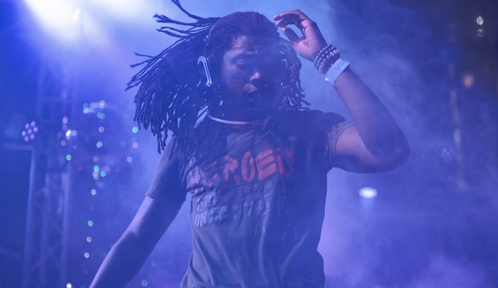 A DJ with long dreadlocks and headphones performs energetically on stage with vibrant lighting.