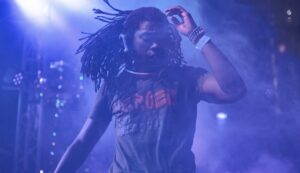 A DJ with long dreadlocks and headphones performs energetically on stage with vibrant lighting.
