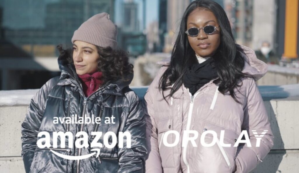 Two people wearing stylish winter jackets stand outdoors in a city setting with the logos "available at Amazon" and "OROLAY" displayed.