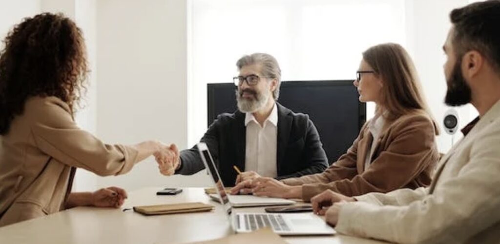 **Alt Text:** Four people are seated around a table in a meeting, with two of them shaking hands.