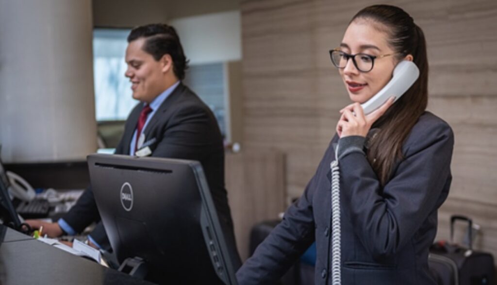 Two hotel receptionists, a man and a woman, are working at a front desk; the woman is speaking on a phone.