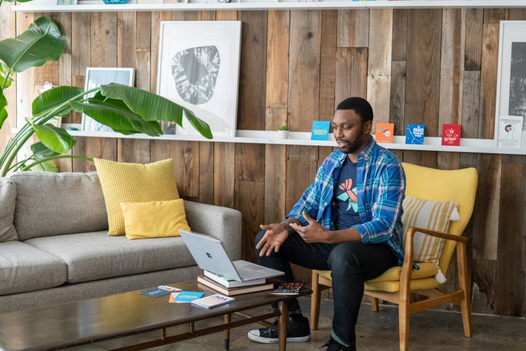 A man sitting in a cozy living room setting, talking to a laptop on a coffee table, surrounded by modern decor and bright cushions.