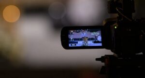 **Alt Text:** A video camera is focused on a stage, capturing a person speaking at a podium.
