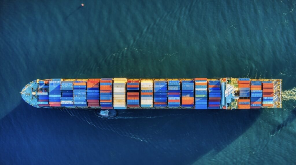 **Alt text:** Aerial view of a large cargo ship carrying colorful containers moving through blue ocean waters.