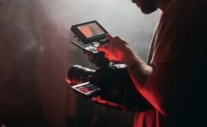 A person is holding a professional video camera with a monitor attached, in low light with a red hue.