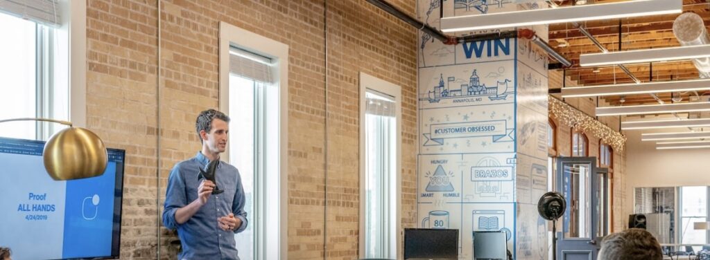 **Alt text:** A man stands in a conference room giving a presentation, with a screen displaying "Proof All Hands 4/24/2019" and a decorated wall with motivational phrases behind him.