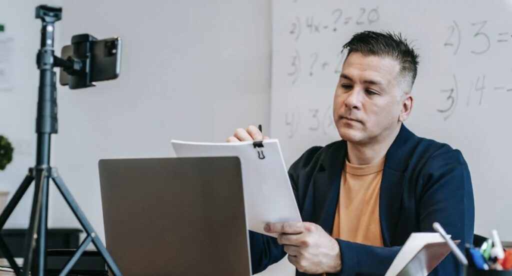 **Alt text:** A man holding papers and looking at a laptop screen is seated in front of a smartphone on a tripod, with a whiteboard featuring math equations in the background.