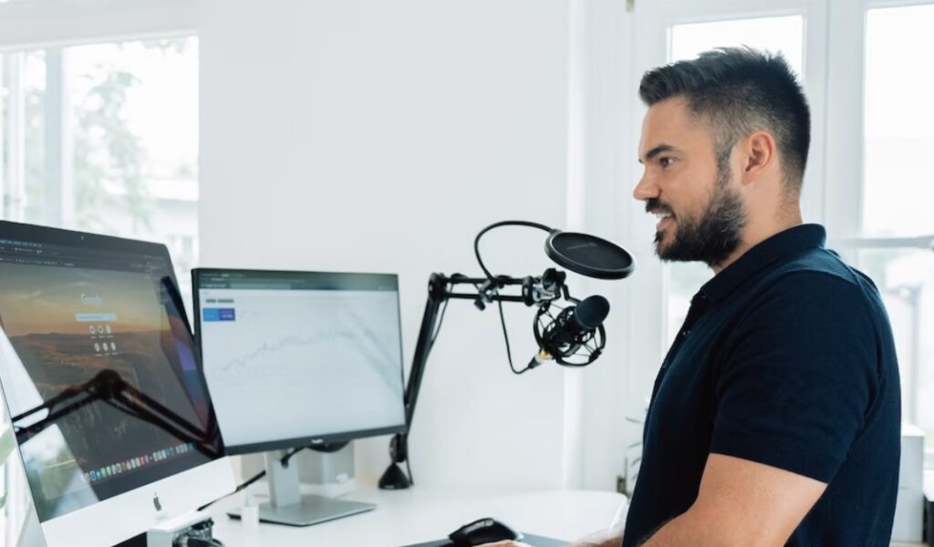 A man standing at a desk with dual monitors and a microphone setup.