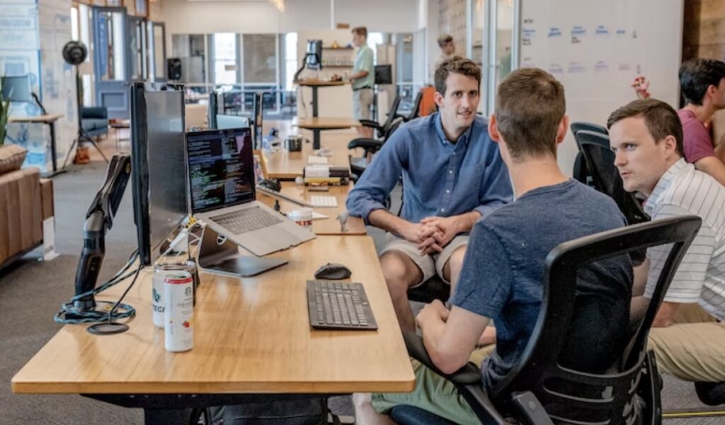 **Alt Text:** Three men sit around a table having a discussion in an open office space with desks, computers, and a laptop displaying code.