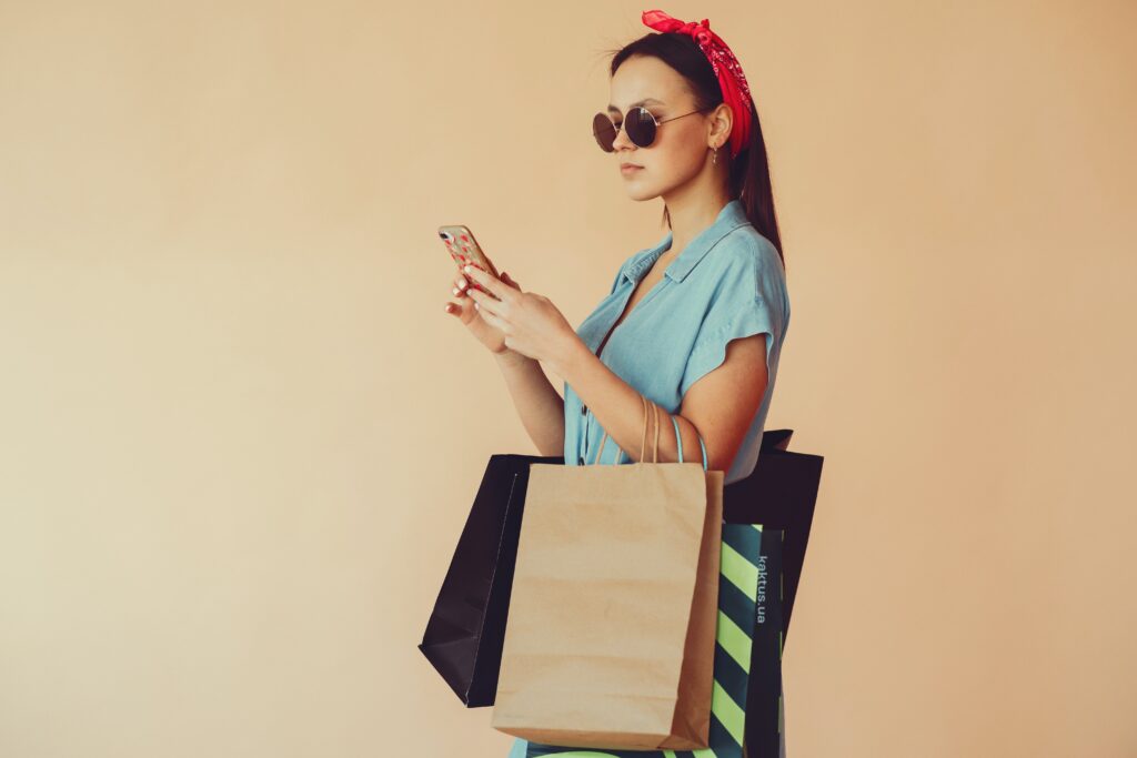 A woman wearing sunglasses and a red headband is holding shopping bags and looking at her phone.