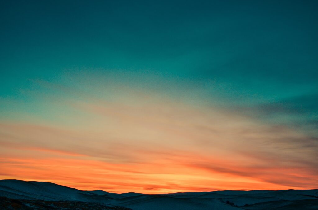 **Alt text:** A panoramic view of a sunset over rolling hills, with a vibrant sky transitioning from deep turquoise to orange and pink hues.