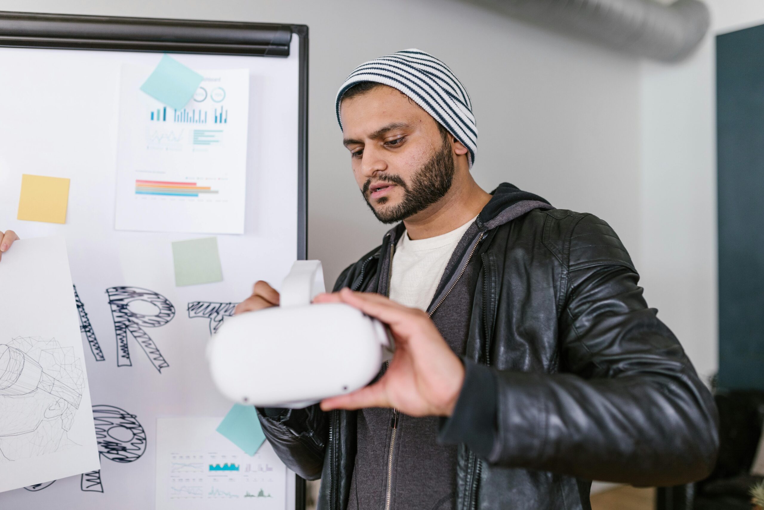 A person wearing a striped beanie and leather jacket is holding a virtual reality headset in front of a whiteboard with graphs and sticky notes.