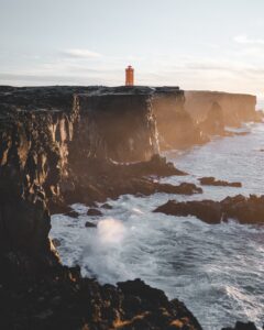 **Alt text**: A bright orange lighthouse stands atop a rugged cliff by a turbulent sea under a clear sky.