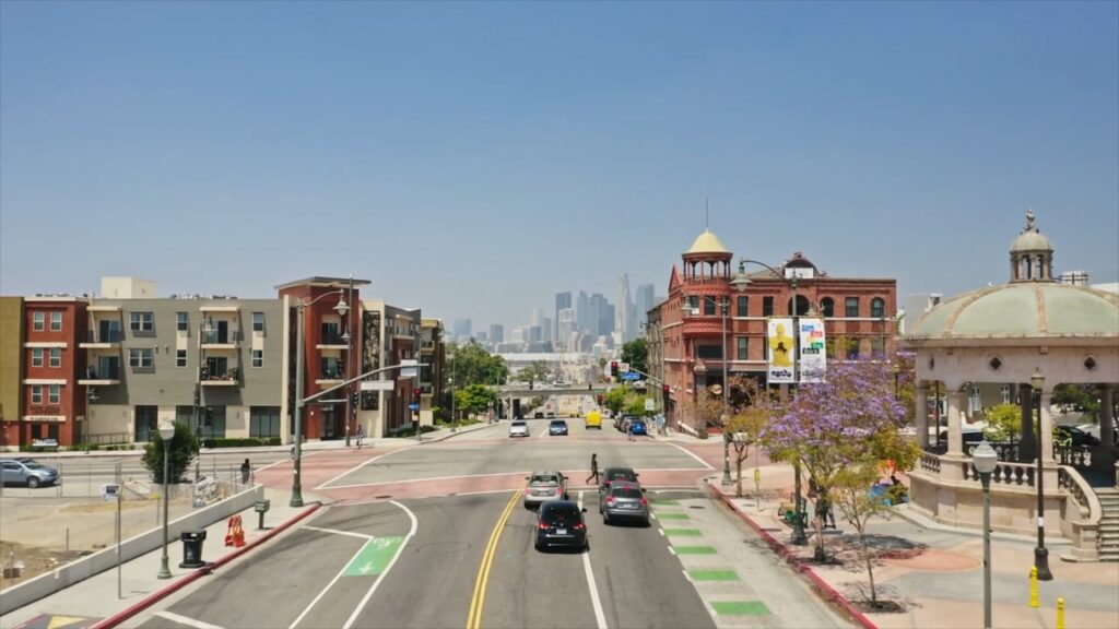 A street lined with modern and historic buildings leads towards a distant city skyline under a clear blue sky.